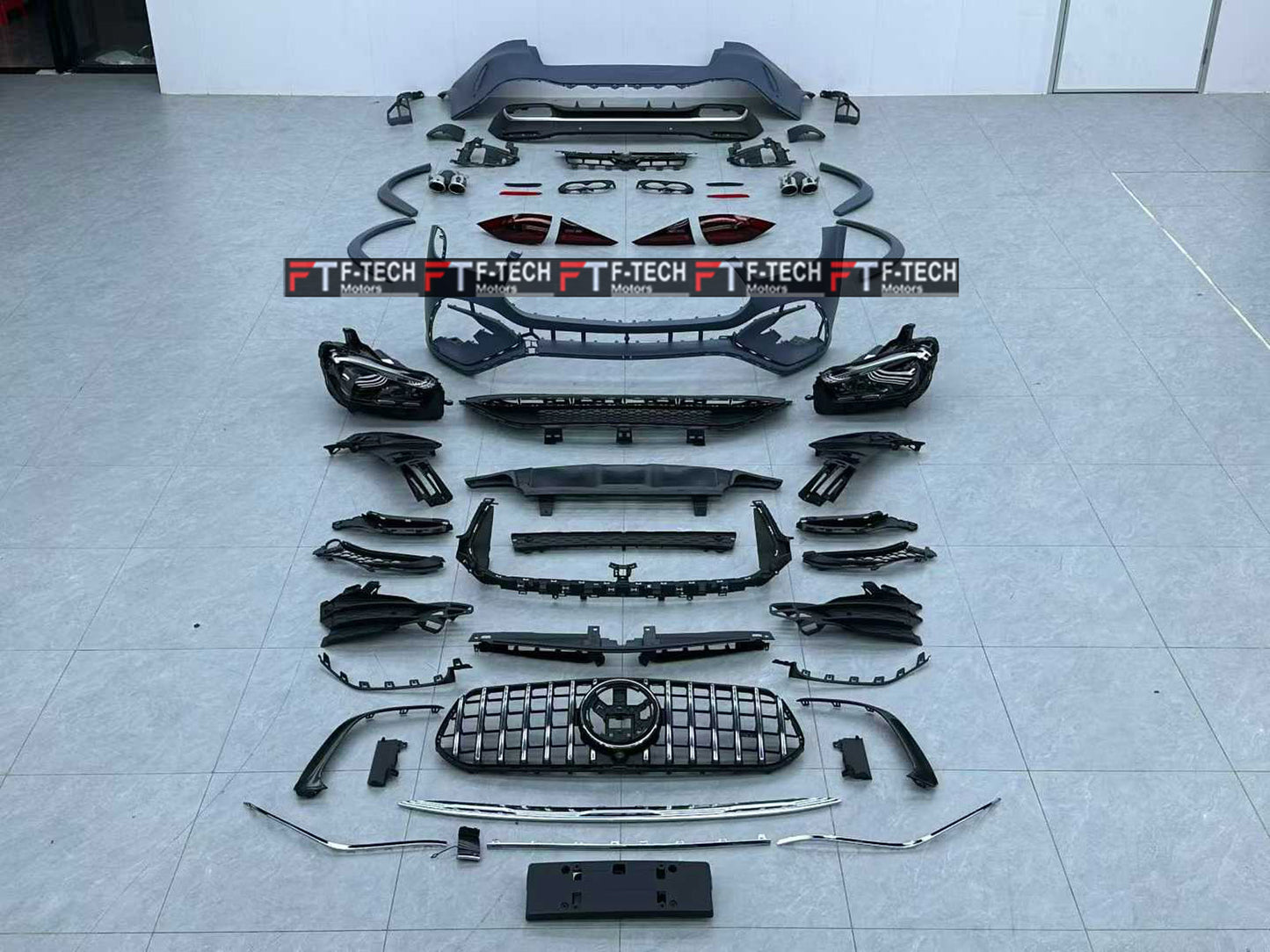 A full set of black car body parts and trim accessories laid out in a row on a light gray tiled floor. The kit includes a front bumper, grille, headlights, fog light housings, side skirts, trim pieces, and other automotive body components.