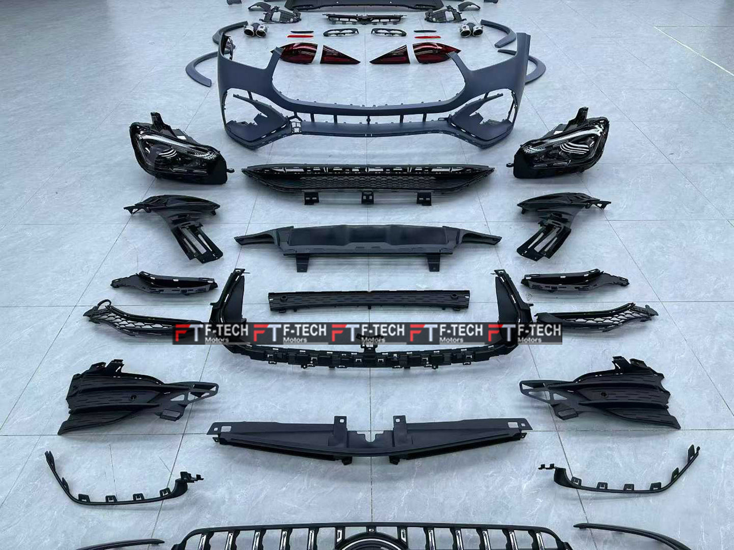 A comprehensive set of black car body parts and grilles, including a front bumper, grille, headlights, fog lights, and various trim pieces, neatly arranged on a light gray tiled floor. A semi-transparent 'T.