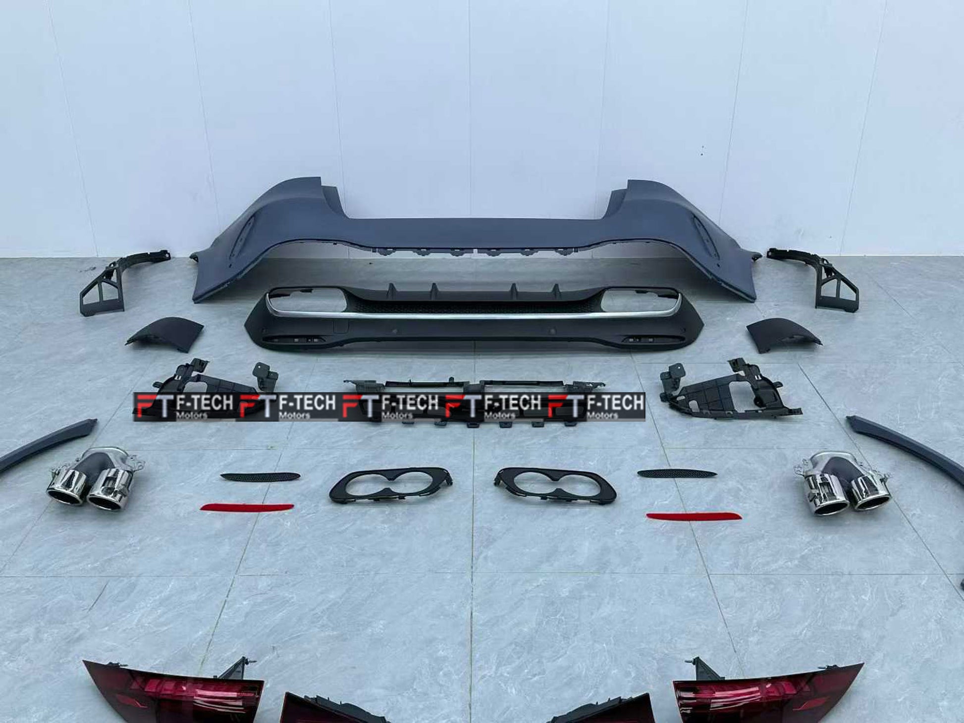A complete car body kit laid out on a light gray tiled floor against a white wall. The kit includes a black rear bumper, a lower lip spoiler, two exhaust tips, two license plate frames with 'F-TECH' branding, trim pieces, and red-tinted tail light covers.