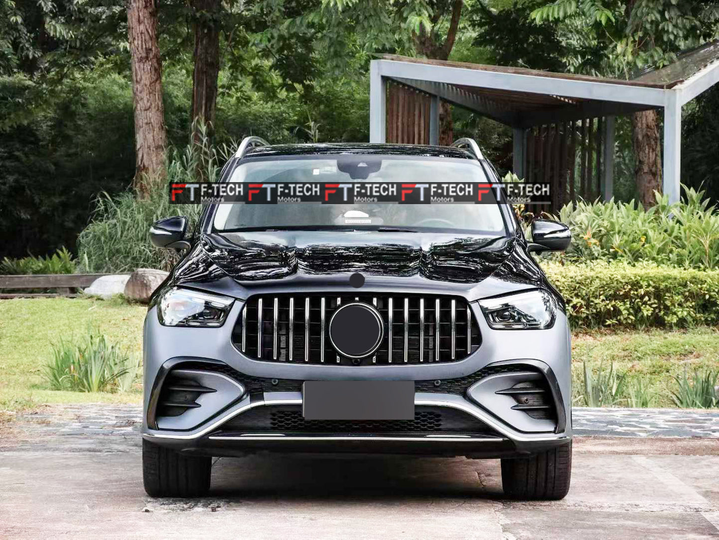 A front-facing view of a dark gray Mercedes-Benz SUV parked outdoors on a paved surface, with trees, grass, and a covered walkway in the background. A repeated 'F-TECH' watermark is visible across the windshield.