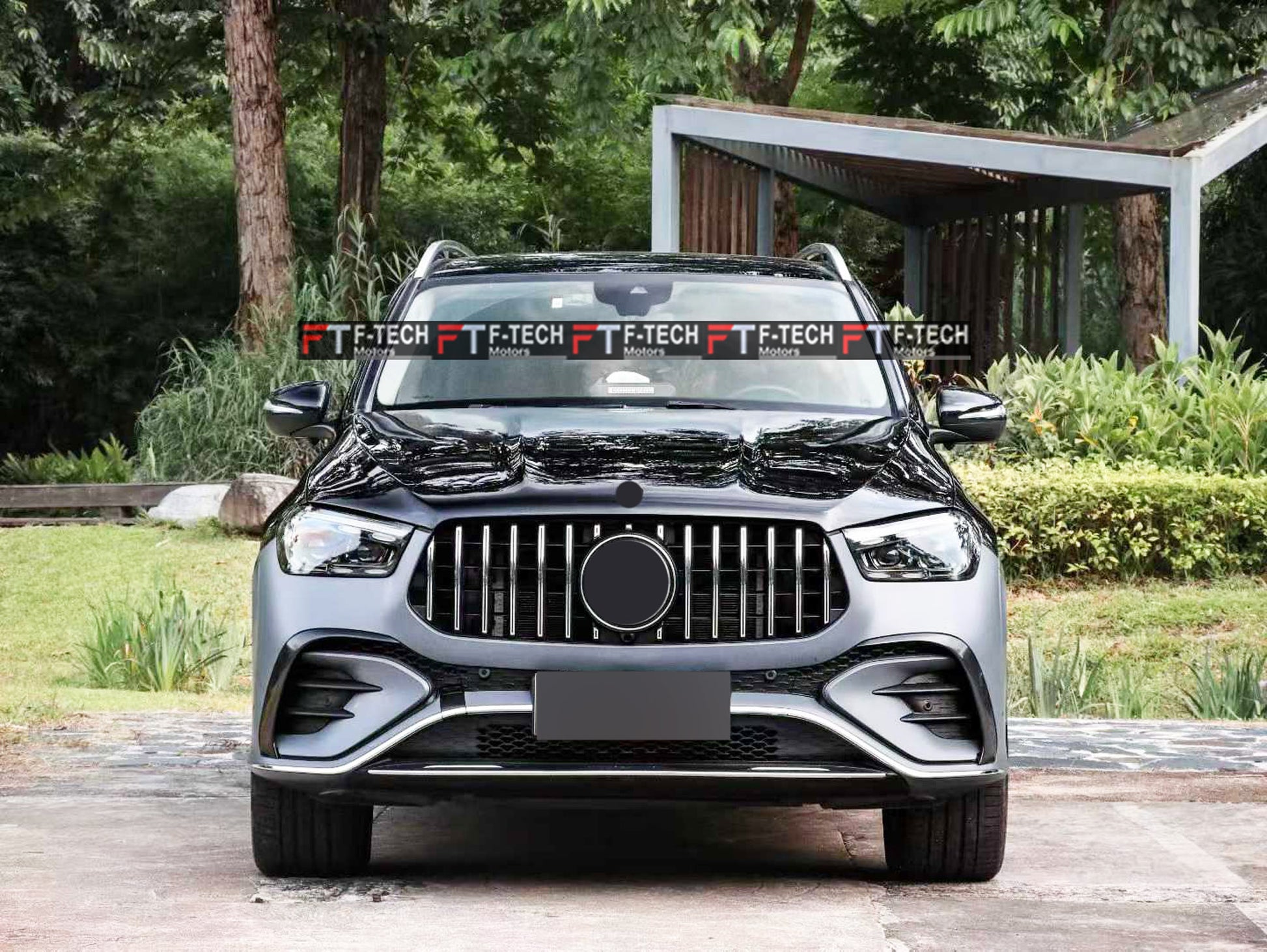 A front-facing view of a dark gray Mercedes-Benz SUV parked outdoors on a paved surface, with trees, grass, and a covered walkway in the background. A repeated 'F-TECH' watermark is visible across the windshield.